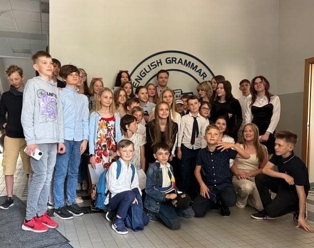 Liela skolēnu un skolotāju grupa pozē skolas vestibilā zem zīmes ENGLISH GRAMMAR