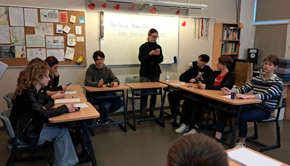 Skolēni klasē sēž pie galdiem un debatē