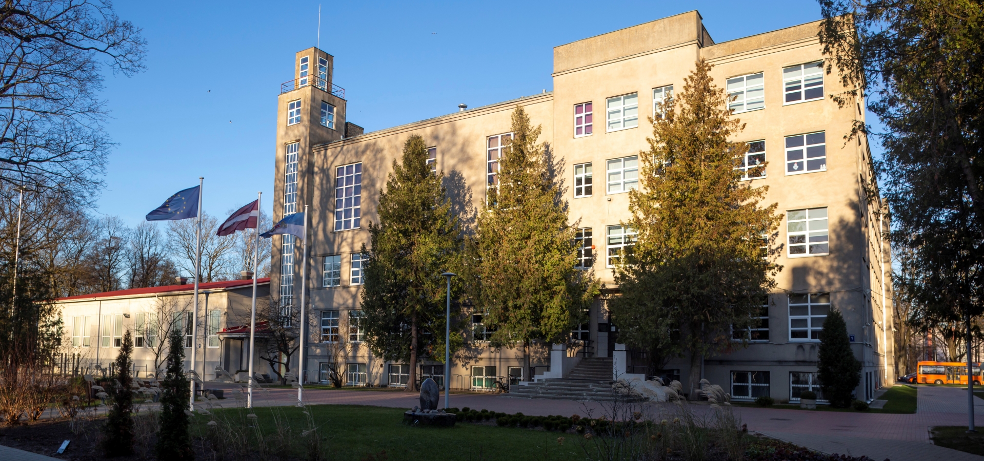 Rīgas Angļu ģimnāzijas ēka ar Eiropas, Latvijas un Rīgas karogiem un kokiem